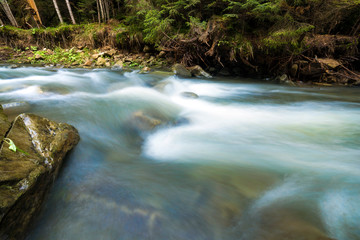 Obraz premium Fast flowing through wild green forest river with crystal clear smooth silky water falling from big wet stones in beautiful waterfalls on bright sunny summer day. Long exposure shot.