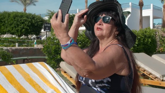 Caucasian senior female elderly in a black hat makes a selfie on a smartphone and sunbathes in the sun while sitting on a sun lounger. Against the backdrop of a tropical landscape, palm trees and
