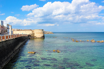 Obraz premium Amazing view of blue sea and adjacent city coast in Syracuse, Sicily, Italy on a sunny day with blue sky. Stones in sea water. Photographed in Ortygia Island. Popular tourist place
