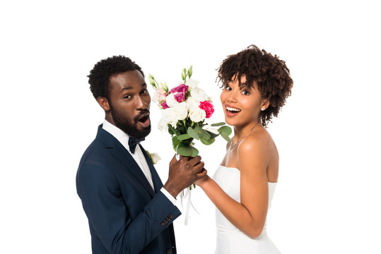surprised african american bridegroom and happy bride holding flowers isolated on white