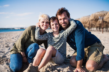 Young family with small boy sitting outdoors on beach, looking at camera.