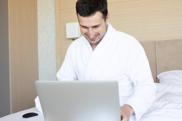 Beautiful young man using laptop while sitting on a bed.