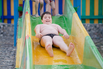 funny delightful fat little caucasian boy in swimming trunks roll down on water slide during summer...