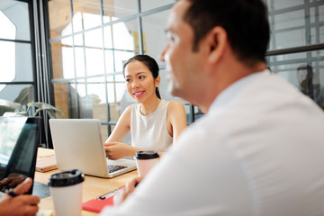 Asian young businesswoman sitting at the table with laptop and smiling while communicating with her colleagues at business meeting at modern office
