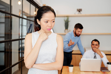 Asian young businesswoman standing at office with thoughtful expression with her colleagues discussing with each other in the background at office
