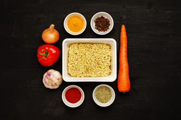 Instant noodles in contaiber with vegetables and spices on a black wooden table, top view