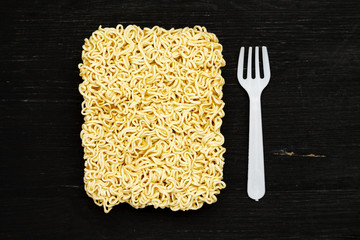 Briquet of instant noodles with plastic fork on a black wooden table, top view