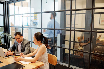 Asian young businesswoman pointing at mobile phone in her hands and discussing with businessman while he working on laptop during a meeting at office