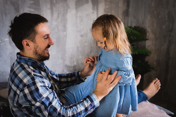 Dad and his daughter play on the bed in the bedroom, smiling and laughing. Daddy's baby. Dad with a beard in a plaid shirt, a girl in a denim dress. Happy family. Father's day