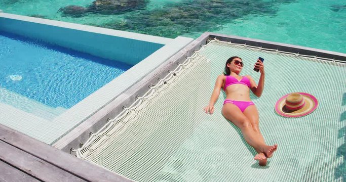 Luxury resort vacation tourist woman relaxing on overwater catamaran net bed in private bungalow suite using phone taking pictures of summer holiday high end hotel.