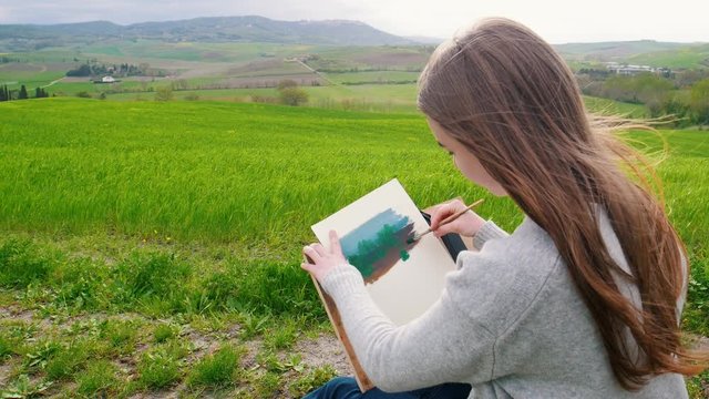 A talanted woman sits on the field and draws using a brush