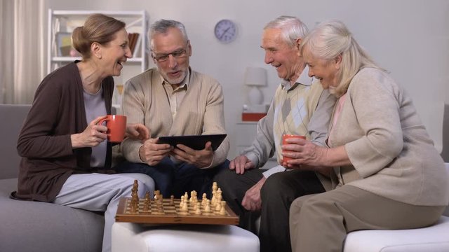 Elderly man showing friends new project on tablet pc, chess play brake, company
