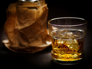 A glass of whiskey with ice on a dark background and a bottle in craft paper.