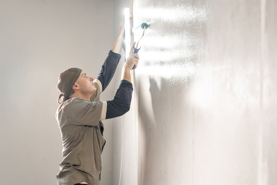 Young repairman smear on wall glue with a roller. . Worker glueing wallpapers on concrete wall. Repair the apartment. Home renovation concept. White Wallpaper for paint