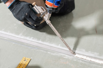 Worker is using a polyurethane foam for gluing drywall at ninety degrees. Hand holding polyurethane expanding foam glue gun applicator - obrazy, fototapety, plakaty