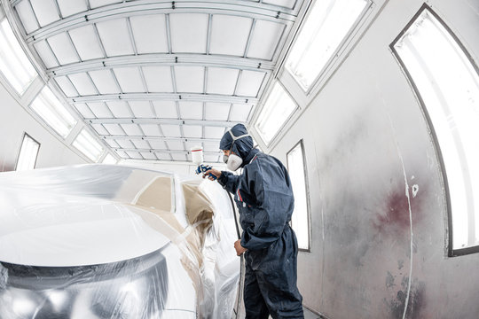 Car service station. Worker painting a white car in special garage, wearing costume and protective gear