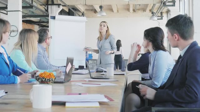 The team leader makes a presentation on a flipchart. Employees sit at the table and discuss. Office life in the background. Coworking. Loft style interior