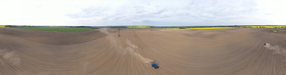 Aerial wide panoramic view on blue tractor pulling a plow, preparing a soil for seed sowing, tractor making dirt cloud. Landmark on meadow and rapeseed field