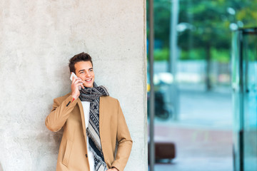 Young man calling. Mid adult man using a mobile phone while standing on grey background. Man leaning on gray wall while looking away and smiling