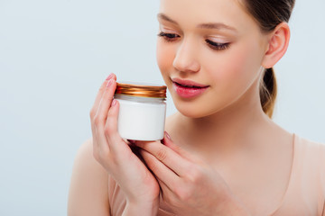 smiling teenage girl looking at container with cosmetic cream isolated on grey