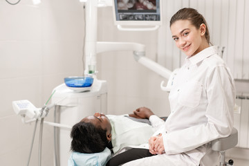 Charming beautiful girl doctor dentist. Young African American male patient at chair at dental clinic. Medicine, health, stomatology concept. dentist conducts inspection and concludes. Woman smiling