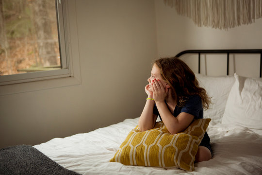 Cute girl with cushion looking through window while sitting on bed at home