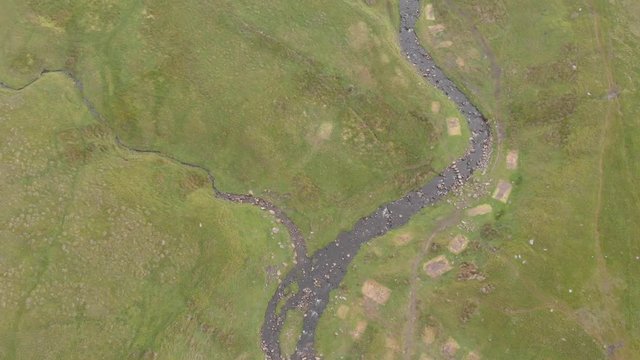 An aerial reveal backward footage of a majestic mountain summit grassy slope with a stream, blue sky and huge altitude white clouds
