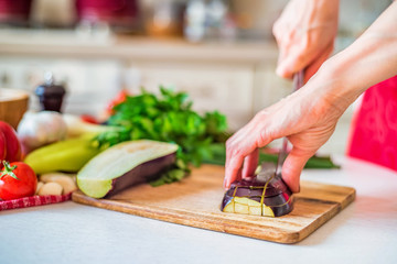 Close up female hand with knife cuts eggplant on wooden board in kitchen