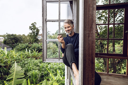Man with cell phone sitting in the window in tropical surrounding