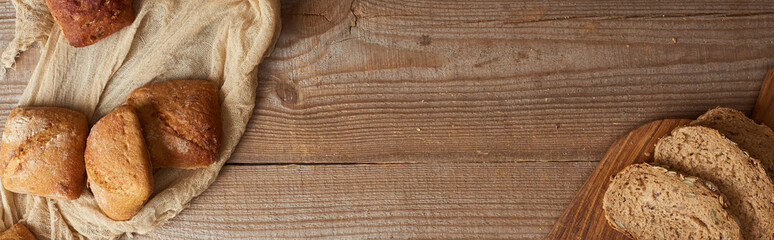 top view of fresh bread slices on chopping board and buns on cloth on wooden table, panoramic shot