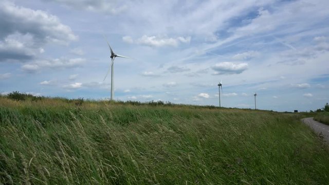 Low angle panoramic view of a wind farm with three wind turbines in rural environment in Zoeterwoude. Windswept grass in the foreground, clouds on blue sky in the background.
