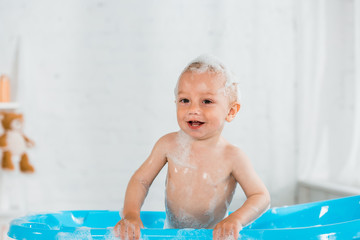 Fototapeta na wymiar adorable toddler kid taking bath and smiling in blue plastic baby bathtub