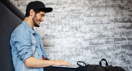 young man in denim jacket and black cap sitting on the floor working on laptop