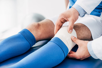 partial view of Physiotherapist putting elastic bandage on leg of football player