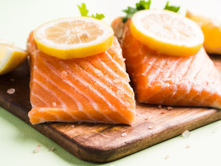 Salmon fillet on wooden board with lemon slices, parsley and pink salt. Pastel green background. Healthy fish meal to be cooked. Protein source.