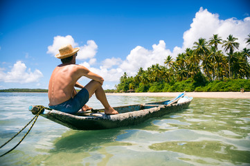 Tourist wearing a straw sun hat sitting in a rustic dugout canoe on a sunny palm-lined tropical...