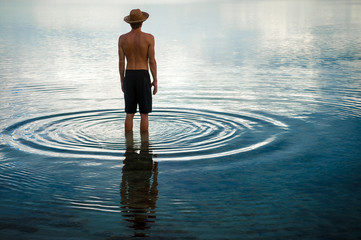Tropical silhouette of man in straw hat standing in calm shallow waters with ripples emanating on the surface - Powered by Adobe
