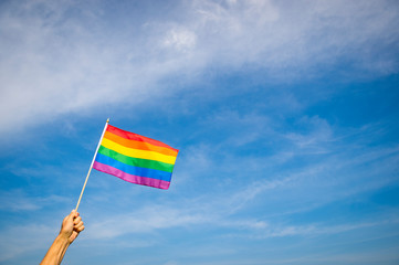 Hand waving gay pride rainbow flag in wide blue summer sky copy space