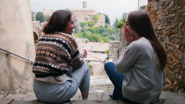 Two young women are sitting on the stairs and talking against the background of buildings