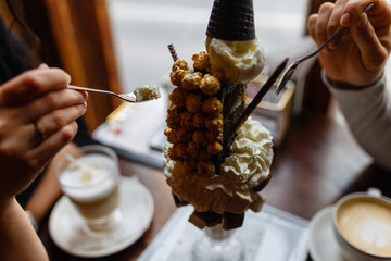 Young couple eating Dessert based on vanilla ice cream and chocolate syrup with cookies, marshmallows, popcorn, cream in glass with at wooden table in cozy cafe. Crazy freakshare food trend.