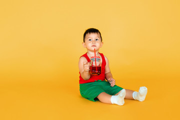 Cute little baby boy in red t-shirt and green short with cup of juice on yellow color background
