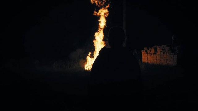 Silhouette of a young woman standing front of a large fire.  Flames and  sparks fly in the background. Slow motion steadicam footage.