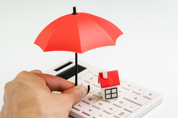 House maintenance and protection and home care service or cost calculation concept, human hand holding small umbrella over group of small miniature houses with red roof on white calculator