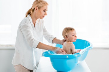 attractive mother washing toddler son in blue plastic baby bathtub