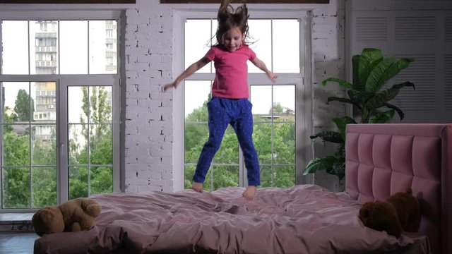 Adorable preschooler girl in pajamas having fun in spacious children's bedroom on background of wide window overlooking green city. Cute elementary age child jumping high on cozy bed in nursery