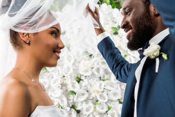 Low angle view of happy African American bridegroom touching white veil and smiling near bride and...