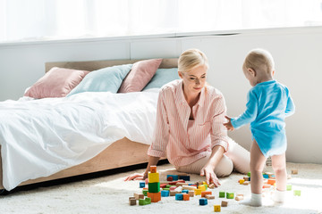 Naklejka premium blonde mother sitting on carpet near toddler son playing with building bricks