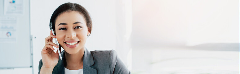 panoramic shot of cheerful latin businesswoman talking on smartphone and smiling at camera