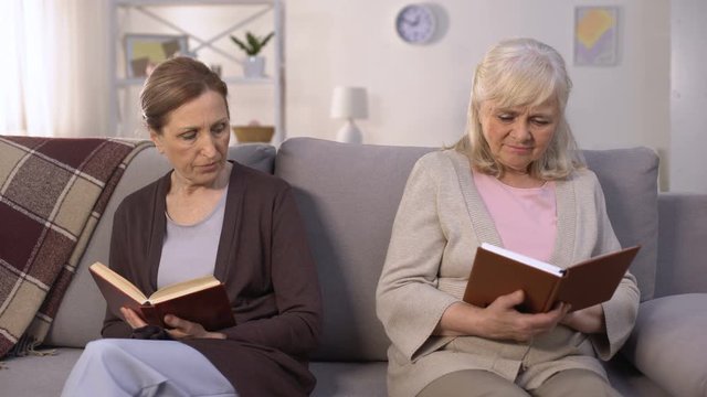 Two envious old women reading books sitting on hall of nursing home, rivalry