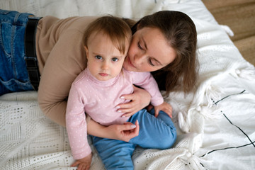 Mom with her daughter lying and resting on the bed. A woman of European appearance, along with a baby in jeans and turtlenecks. Relaxing on the weekend.